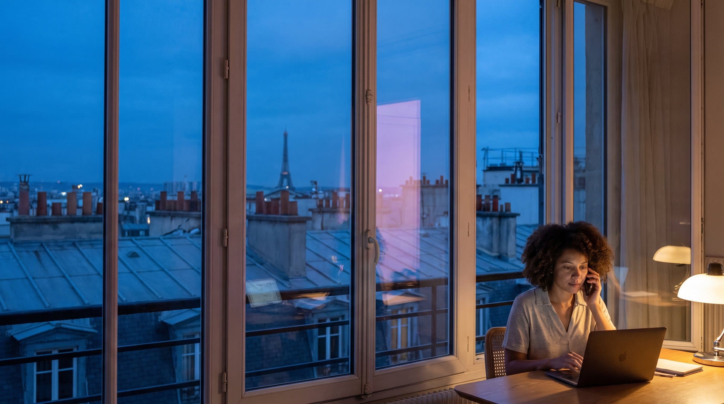 Toits de Paris à l’heure bleue, la tour Eiffel au loin, une femme au téléphone derrière une fenêtre éclairée